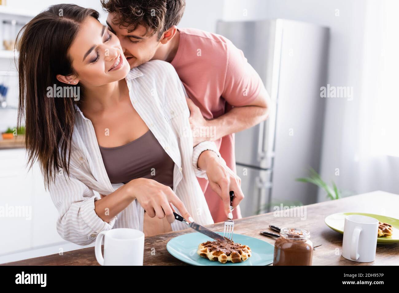 Uomo baciante ragazza sorridente tagliando cialda vicino al cioccolato spalmato e. tazze sul tavolo Foto Stock