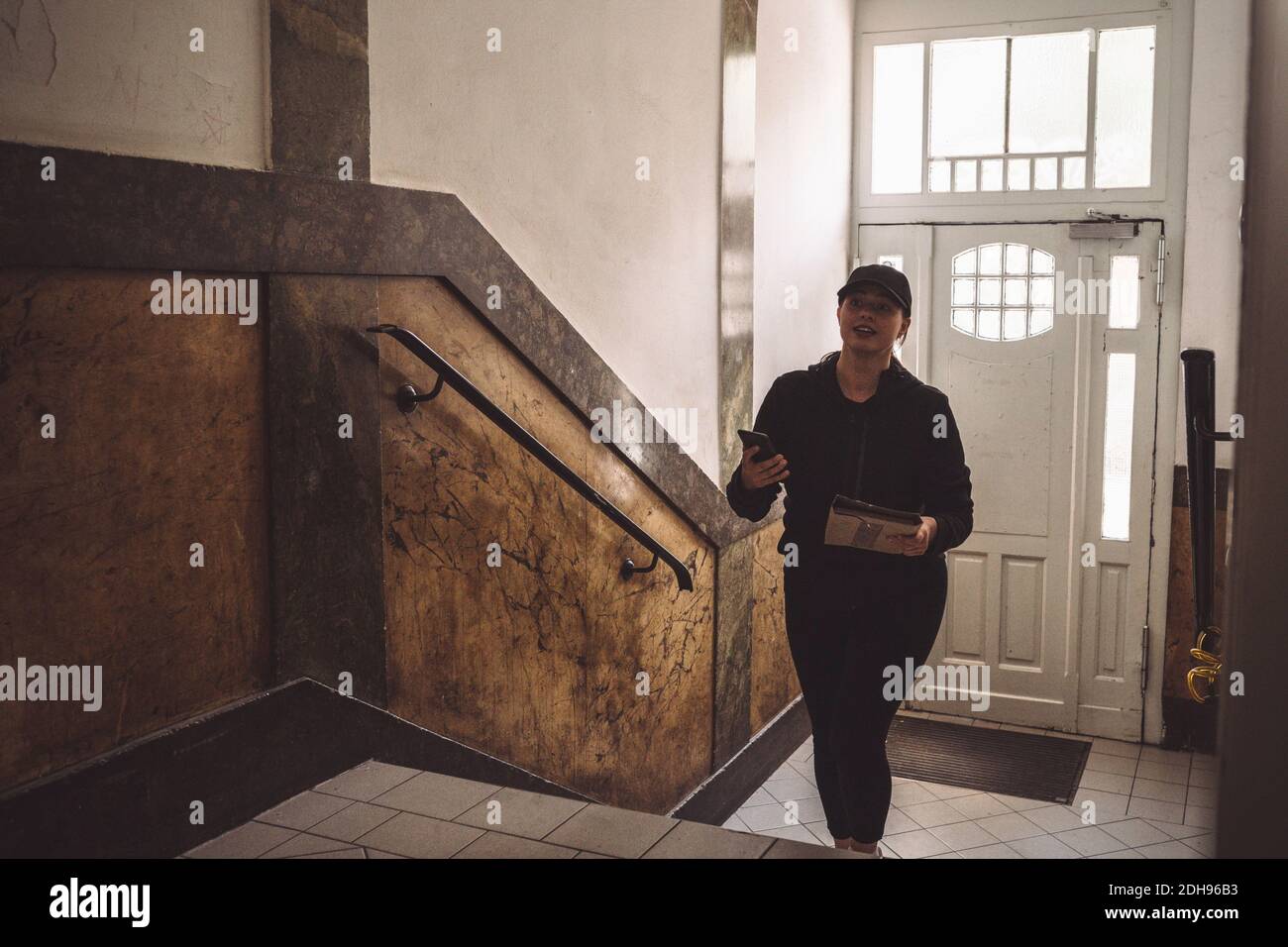 Donna sicura di consegna con telefono e pacchetto salendo le scale contro porta Foto Stock