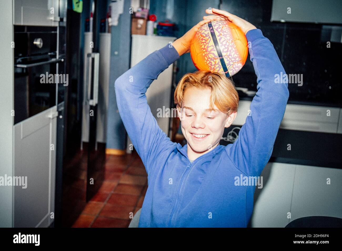 Ragazzo sorridente che tiene la palla sportiva sulla testa in cucina Foto Stock