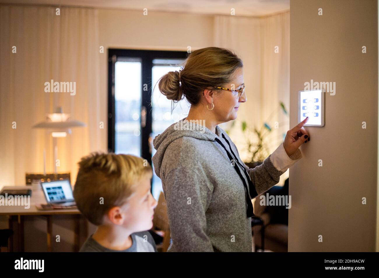 Figlio che guarda la madre utilizzando un tablet digitale montato a parete a casa intelligente Foto Stock