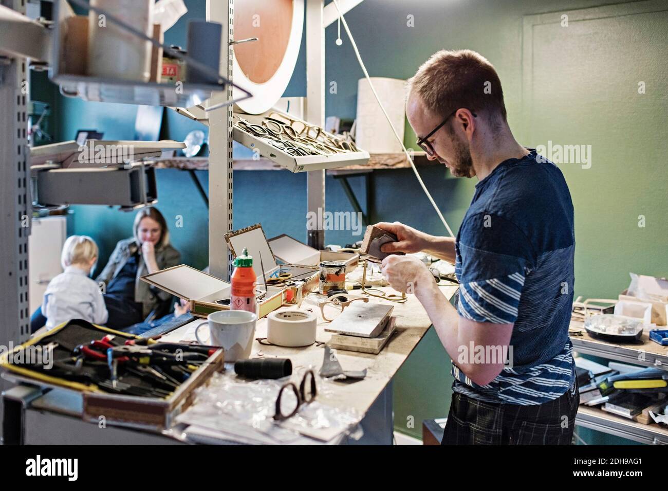 Il proprietario maschile lavora alla scrivania in un'officina di occhiali mentre è in famiglia seduta in background Foto Stock