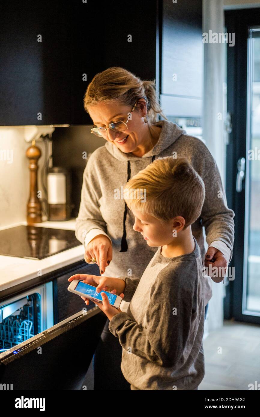 Madre guida il figlio mentre usa l'app mobile in lavastoviglie cucina a casa Foto Stock