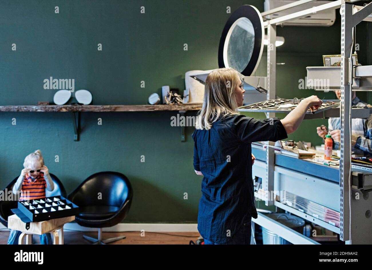 Donna che lavora in laboratorio di occhiali con figlio seduto in background Foto Stock