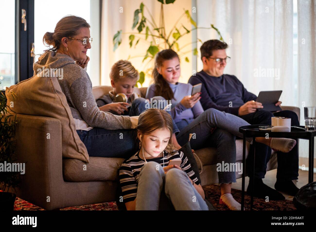 I genitori si siedono con i bambini mentre usano le tecnologie wireless nella vita camera in casa moderna Foto Stock