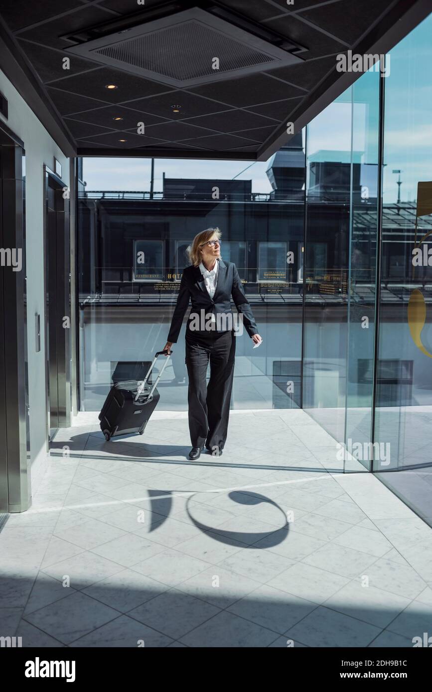 Donna d'affari matura che tira i bagagli su ruote nel corridoio dell'hotel Foto Stock