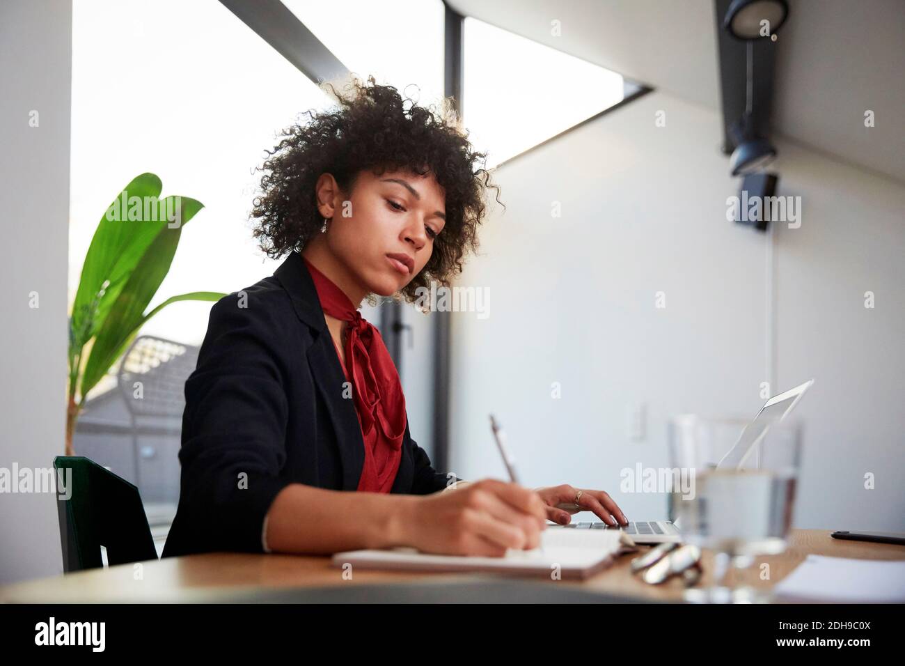 Giovane consulente finanziario sicuro che scrive sul diario mentre sedendo con computer portatile alla scrivania in ufficio Foto Stock