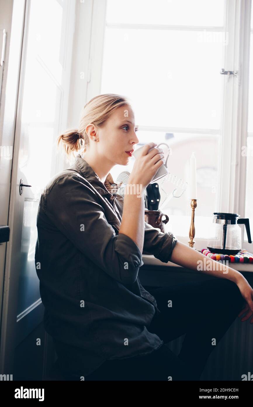 Vista laterale della donna che beve caffè contro la finestra in cucina a casa Foto Stock
