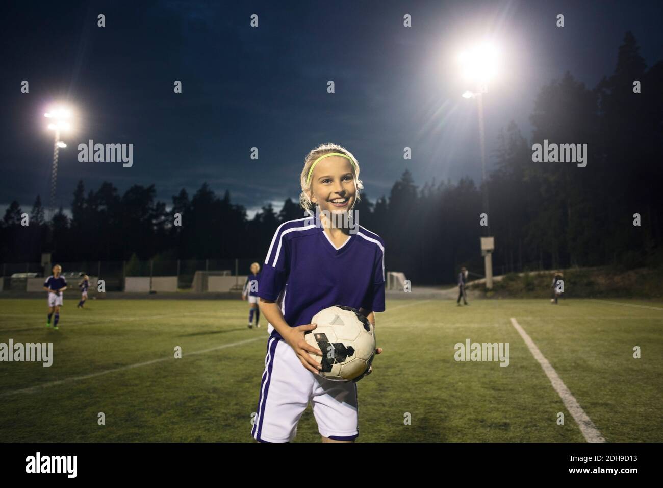 Felice di ragazza in piedi con la palla di calcio sul campo contro cielo di notte Foto Stock