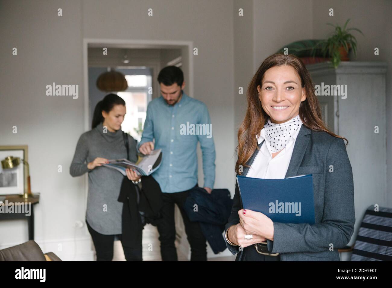 Ritratto di sorridente maturo realtor donna in piedi con coppia di discutere in background Foto Stock