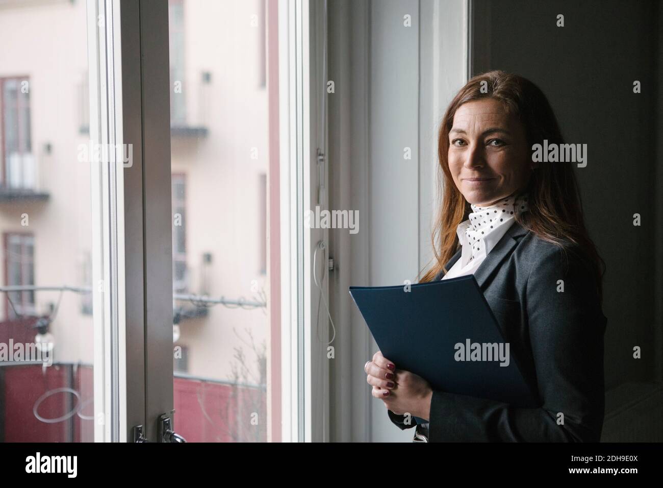 Ritratto di sicuro maturo agente immobiliare in piedi con brochure per finestra a casa Foto Stock
