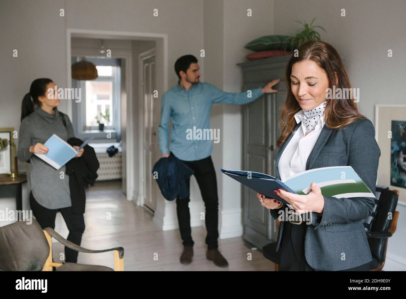 Sorridente agente immobiliare donna che legge brochure mentre coppia cerca a casa Foto Stock