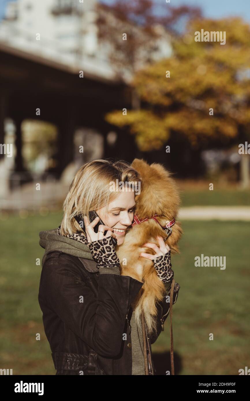 Donna bionda sorridente che parla sul telefono cellulare mentre trasporta Pomeranian al parco il giorno di sole Foto Stock