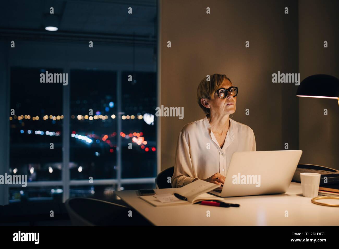 Donna d'affari premurosa seduta con computer portatile alla scrivania illuminata in creativo ufficio Foto Stock