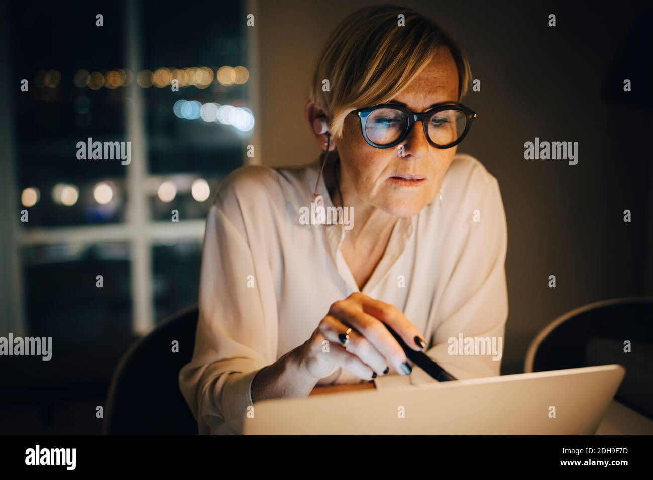 Donna professionale matura dedicata che utilizza un computer portatile in un ufficio illuminato mentre lavoro in ritardo Foto Stock
