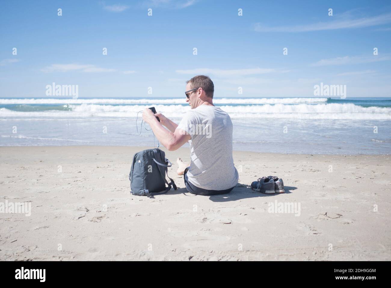 Vista posteriore dell'uomo con telefono cellulare mentre si prendono gli auricolari fuori dello zaino in spiaggia Foto Stock