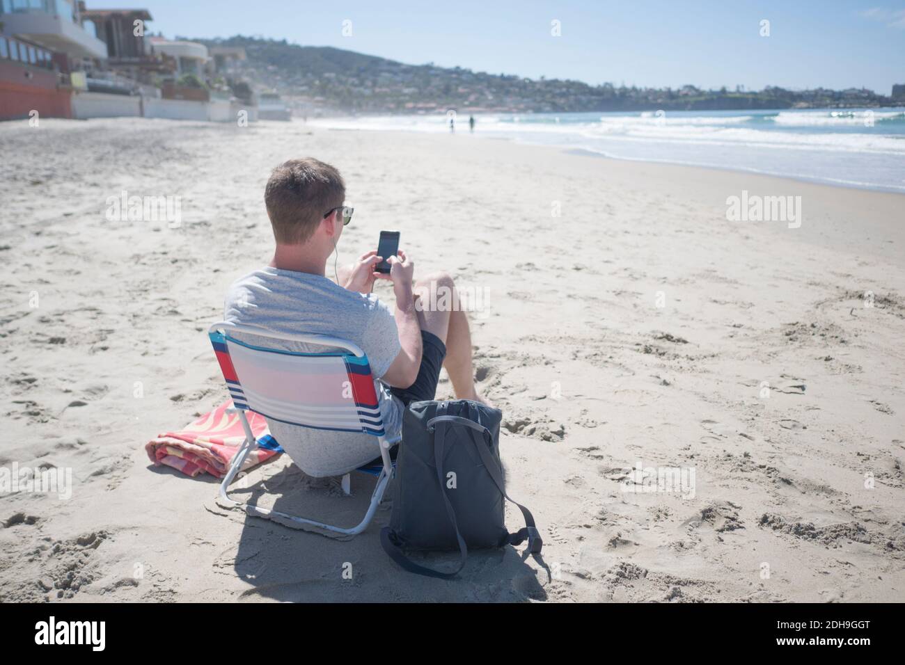 Vista posteriore di un uomo che utilizza lo smartphone mentre è seduto sedia in spiaggia Foto Stock