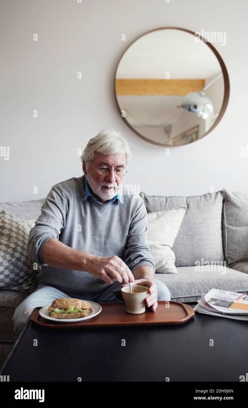 Uomo anziano che mescola il tè mentre fa colazione al tavolo soggiorno Foto Stock