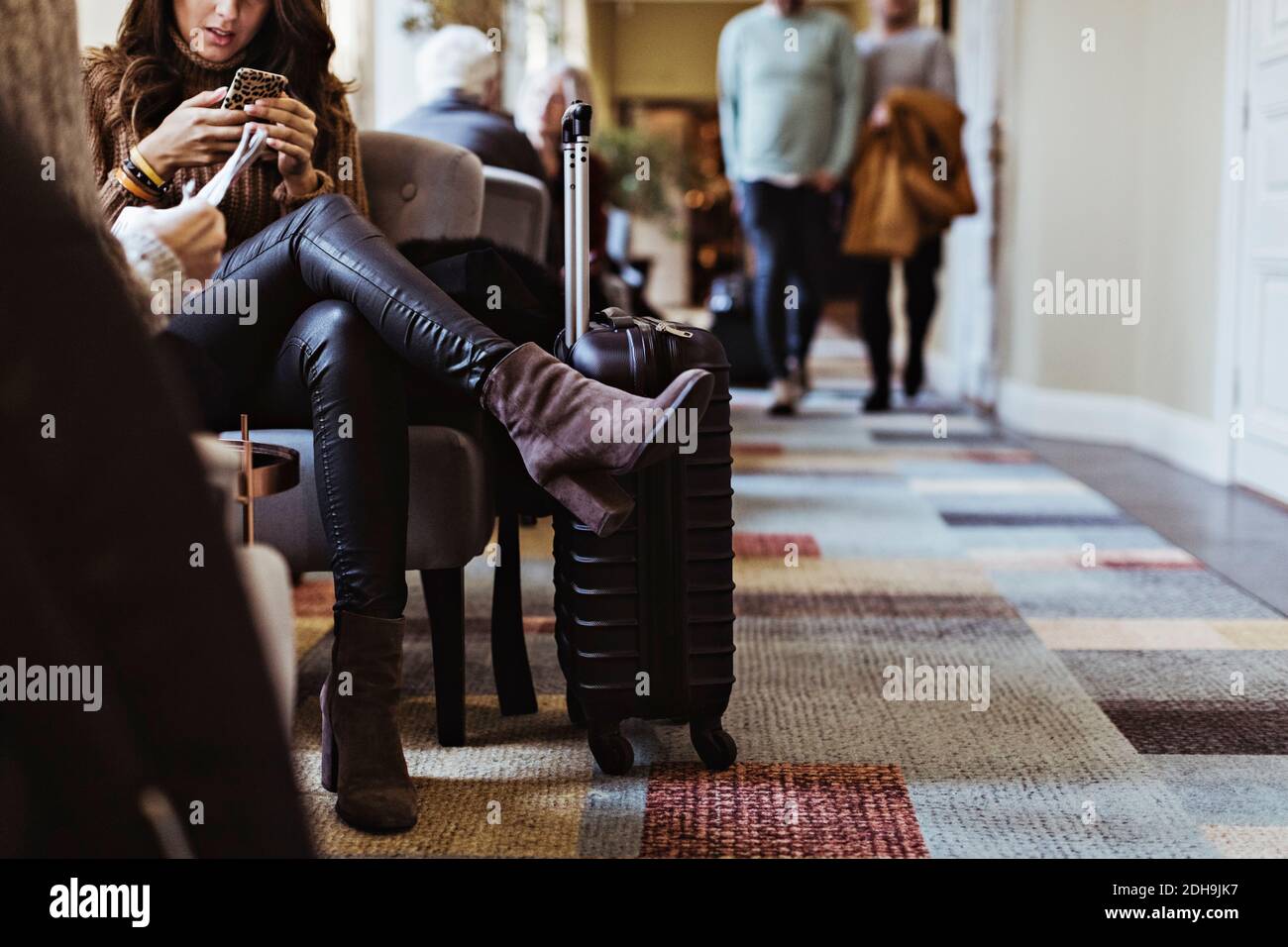 Sezione bassa della donna dell'adulto medio che usa il telefono mobile mentre sedersi sulla sedia nella hall dell'hotel Foto Stock