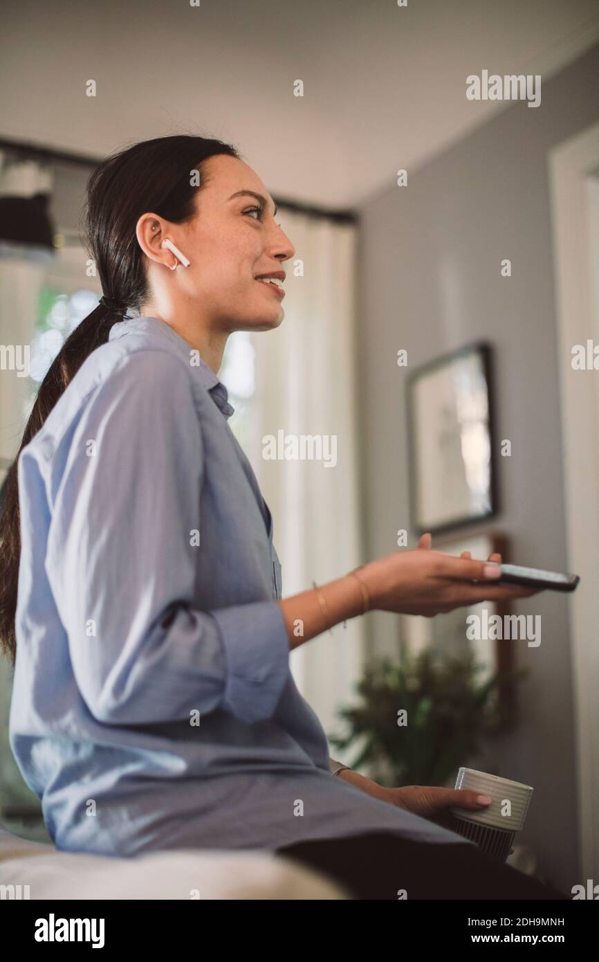 Professionista sorridente con la tazza e il telefono cellulare che parlano attraverso gli auricolari in ufficio a casa Foto Stock