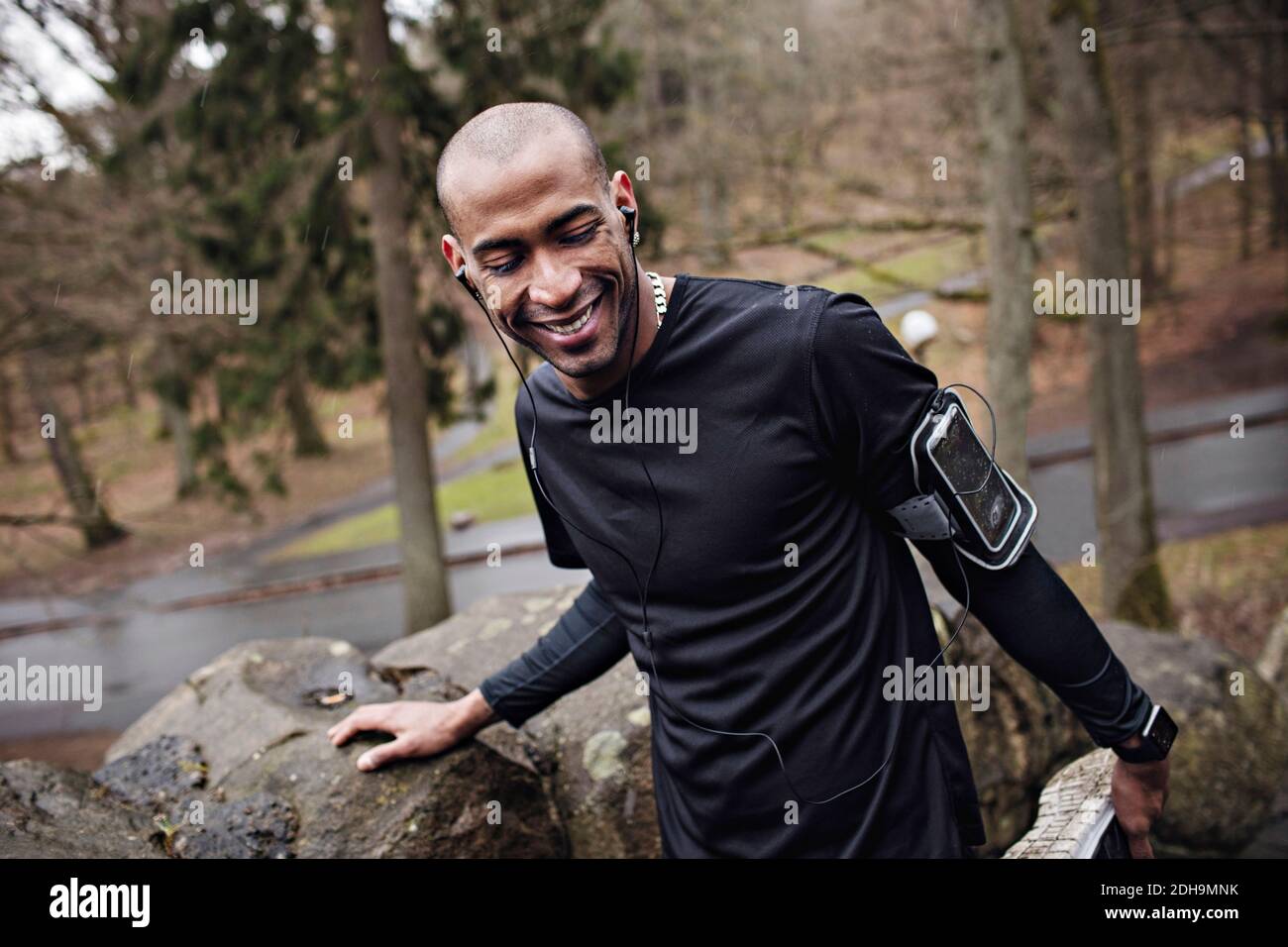 Felice atleta maschile ascoltando musica mentre si esercita da muro di pietra in foresta Foto Stock