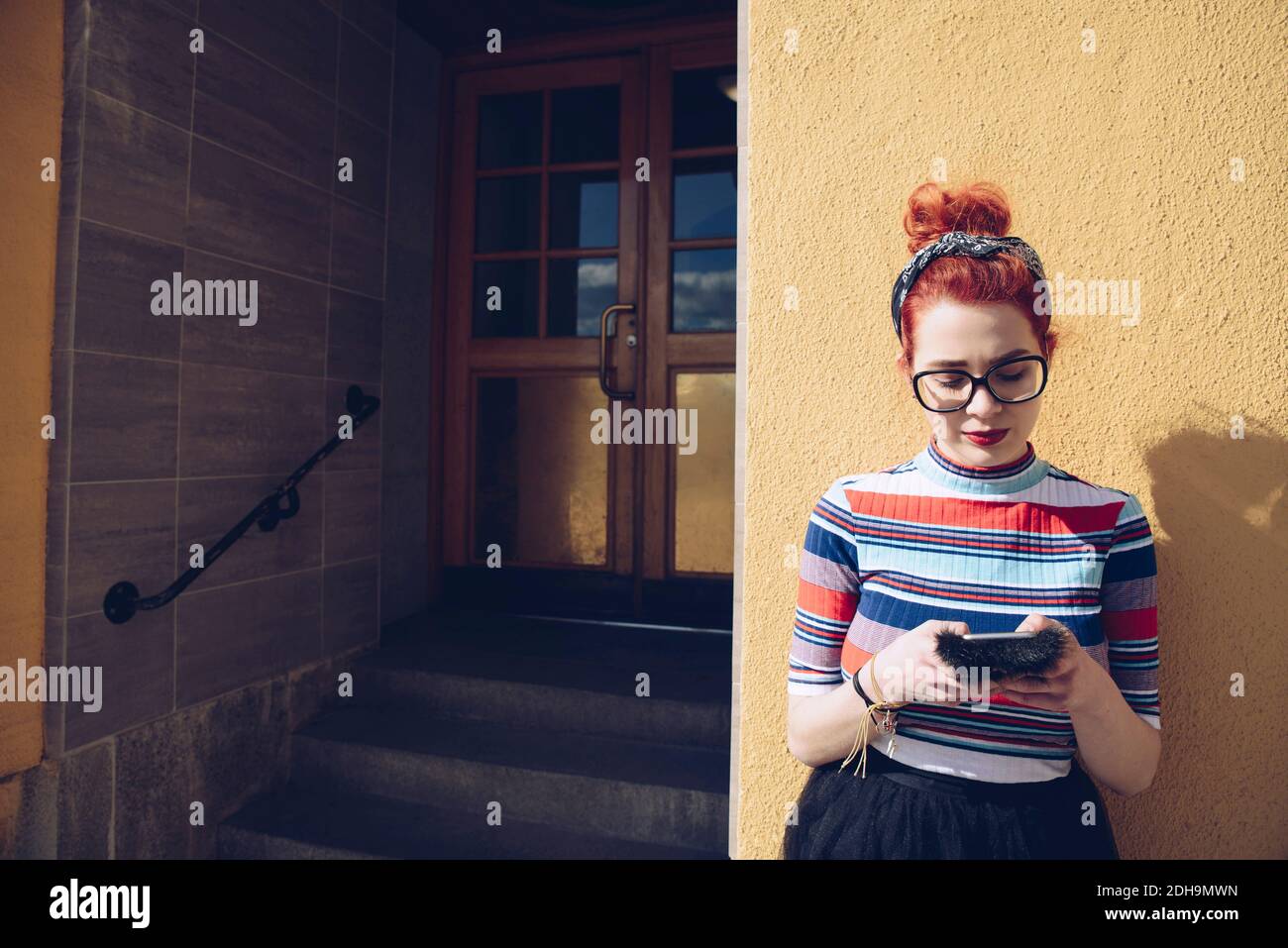 Giovane hipster femminile che usa il telefono cellulare mentre si trova in piedi contro la parete all'ingresso dell'edificio Foto Stock