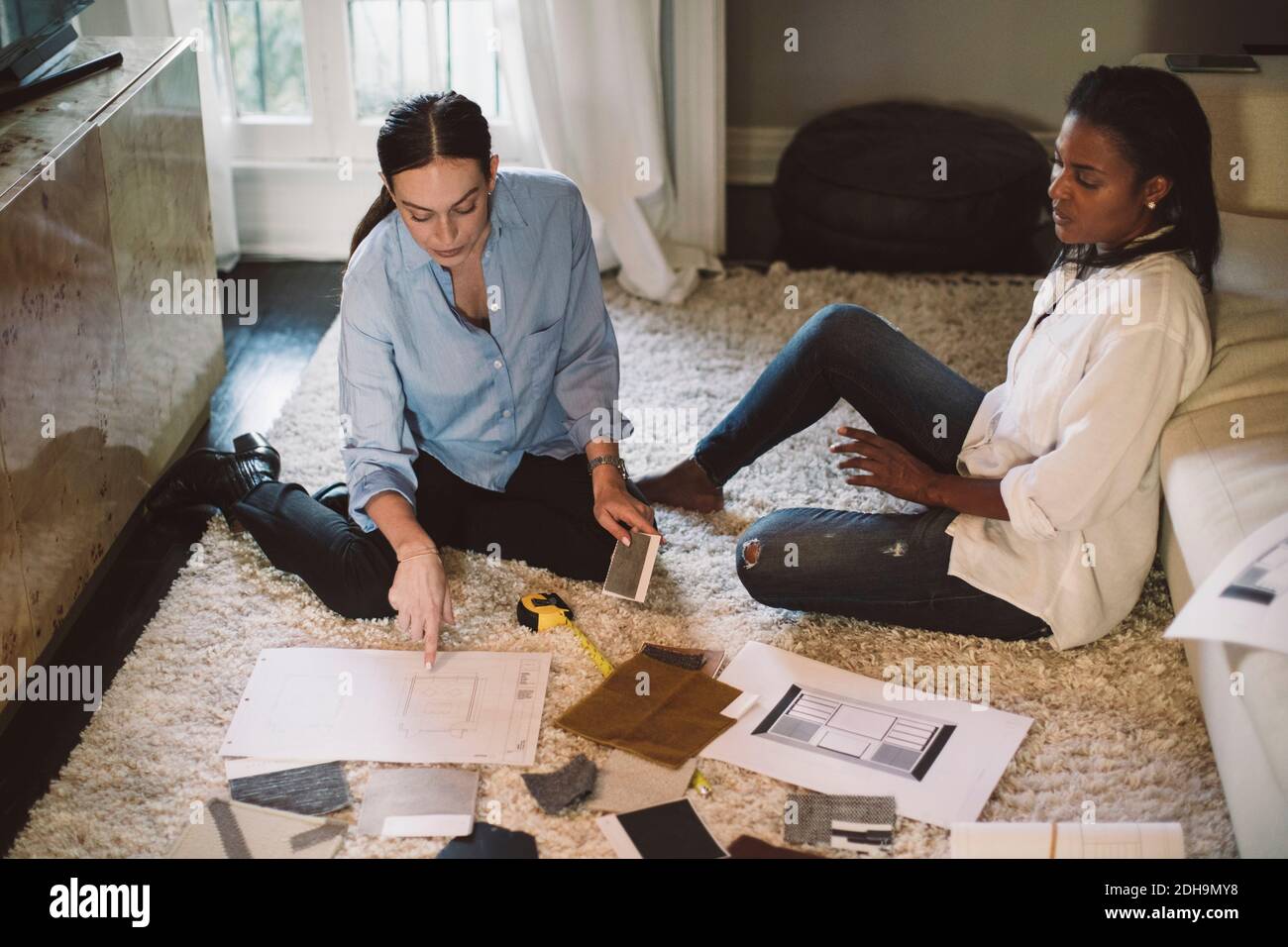 Vista ad alto angolo delle designer femminili che discutono del campione di tessuto mentre si siede sul tappeto in ufficio domestico Foto Stock