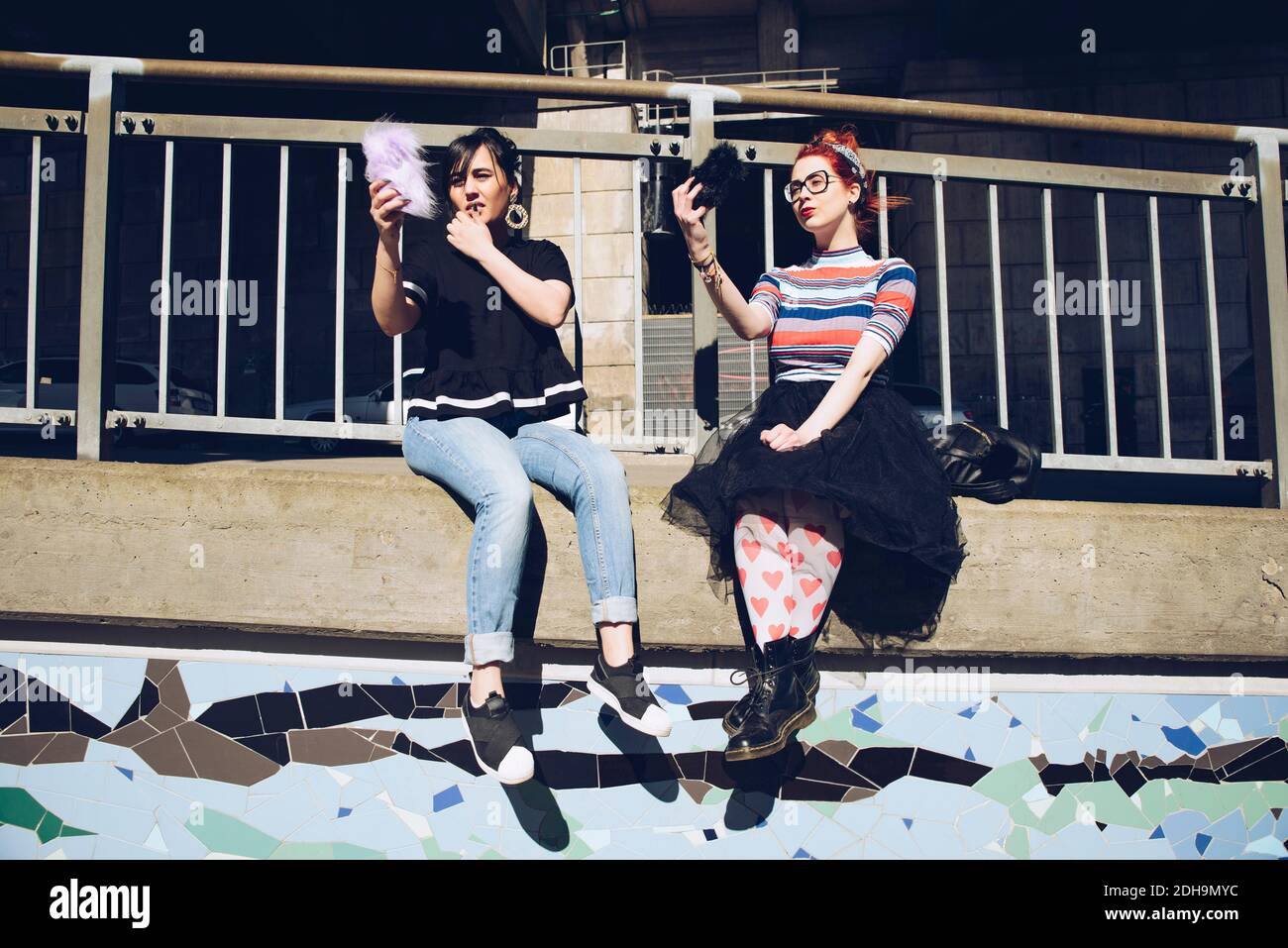 Hipster femmina amici che prendono selfie con i telefoni cellulari mentre si siede su parete contro ringhiera Foto Stock