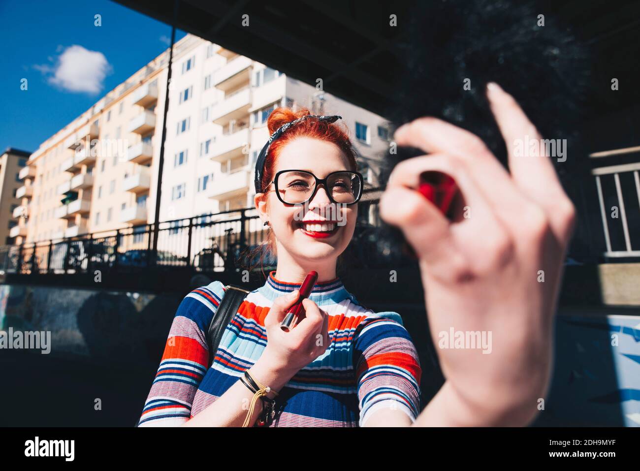 Giovane donna che guarda il telefono cellulare mentre si applica il rossetto città Foto Stock