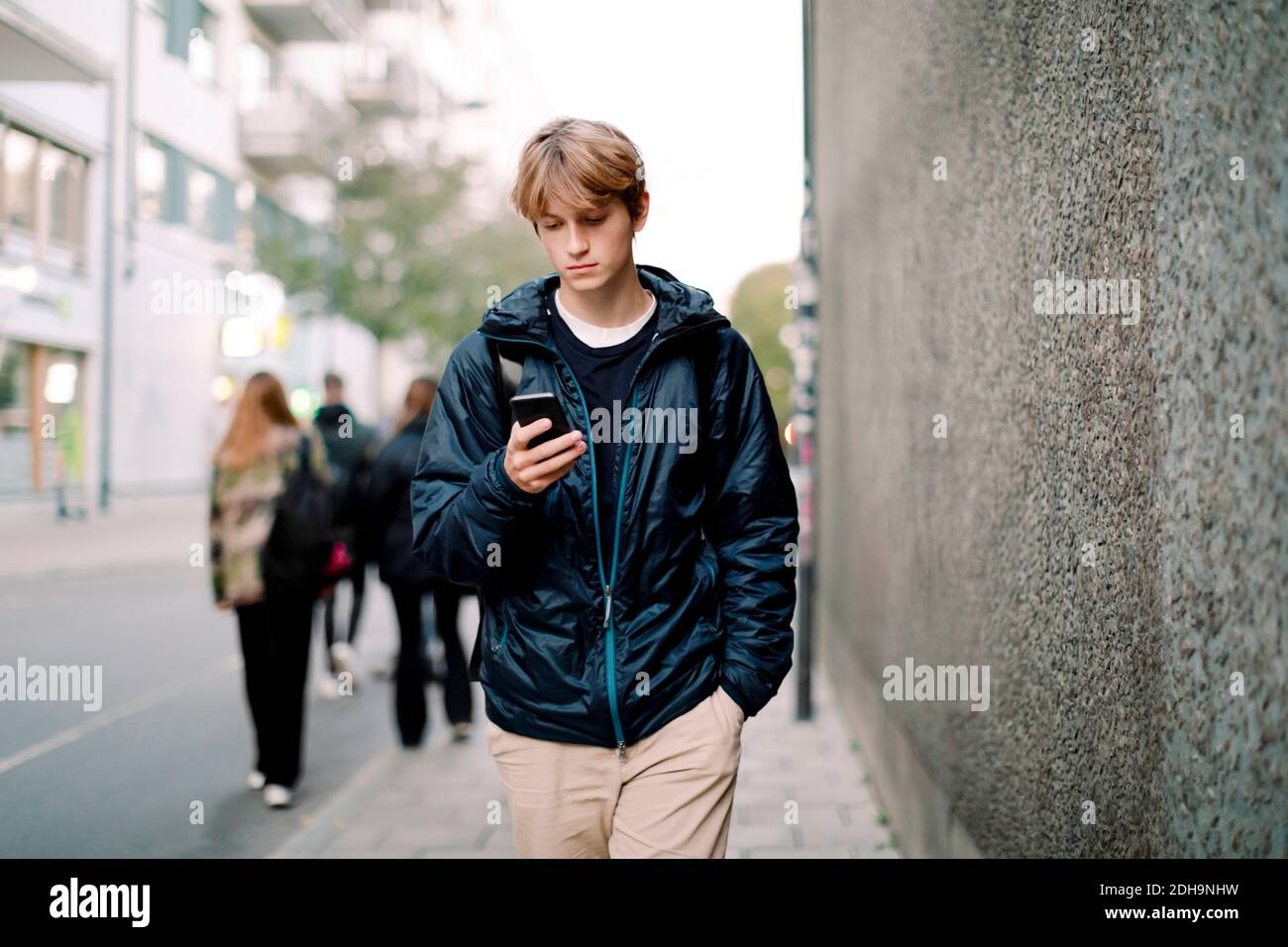 Ragazzo adolescente con capelli biondi che usa lo smartphone mentre cammina sul marciapiede contro le mura della città Foto Stock