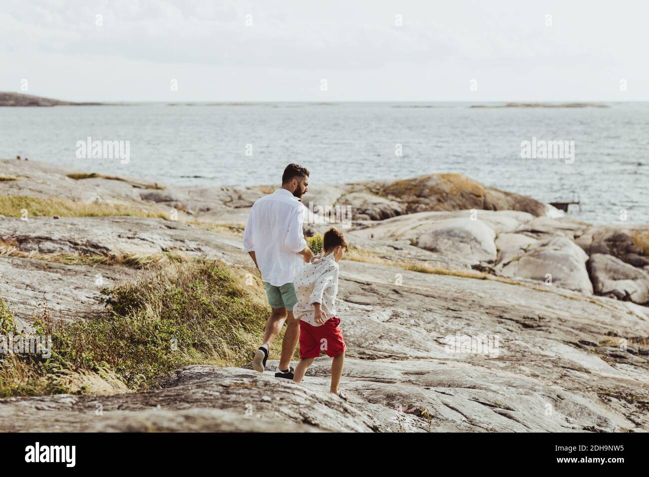 Vista posteriore di padre e figlio che camminano verso il mare terreno roccioso Foto Stock