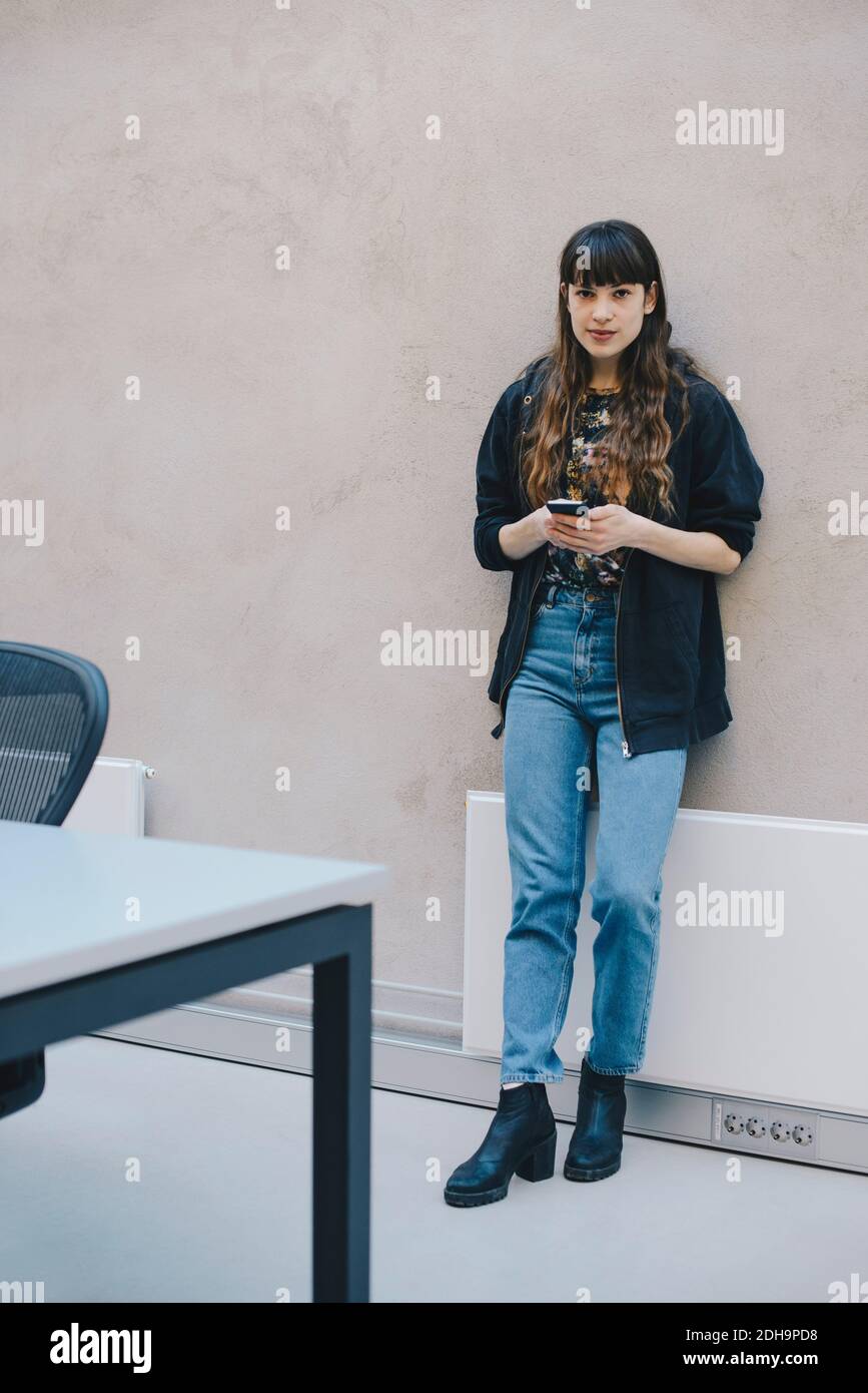Ritratto a tutta lunghezza di una programmatore di computer femminile che tiene uno smartphone mentre si è in piedi contro il muro beige in ufficio Foto Stock
