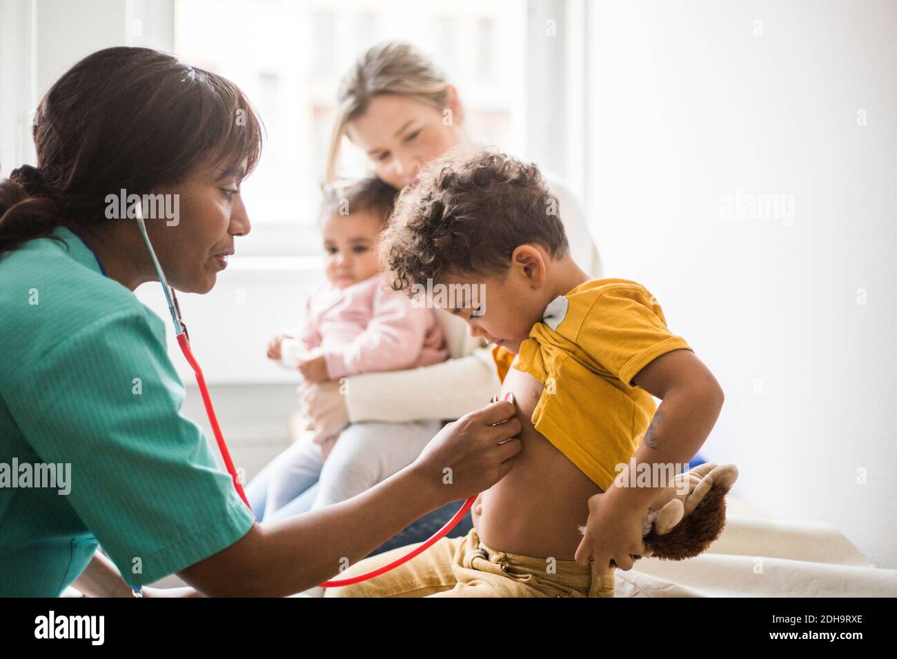 Vista laterale del medico femminile che esamina il battito cardiaco del ragazzo con stetoscopio mentre madre seduta e figlia in clinica Foto Stock