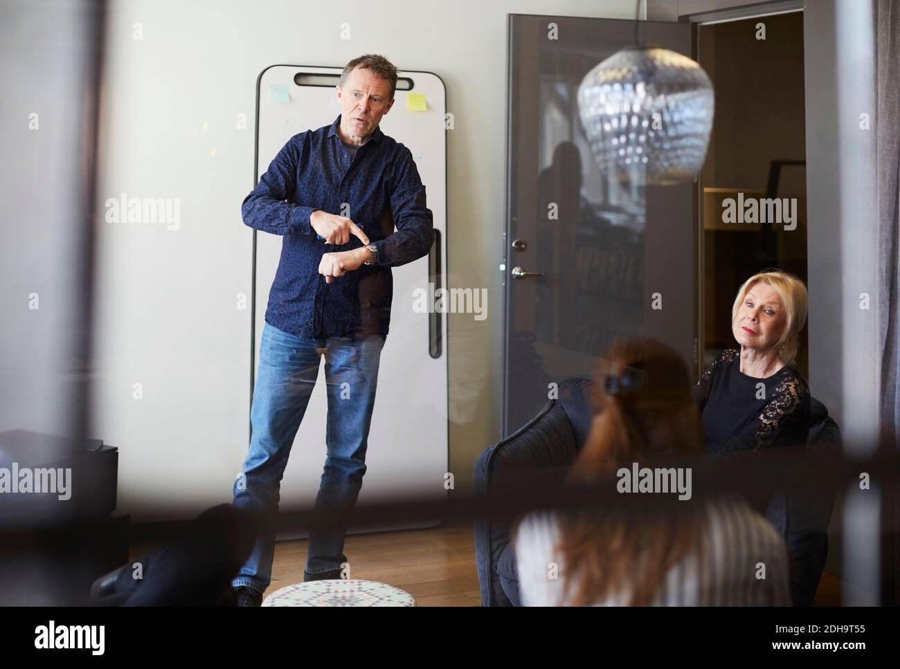 Uomo d'affari che punta al orologio da polso mentre si trova in piedi nella sala da tavola con colleghi di sesso femminile visti dal vetro Foto Stock