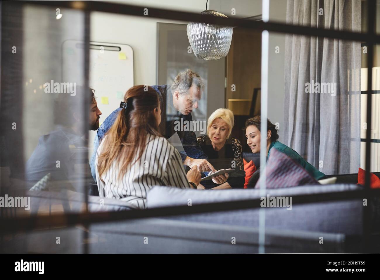Uomo d'affari che mostra il tablet digitale ai colleghi nella sala riunioni durante incontro visto dal vetro Foto Stock