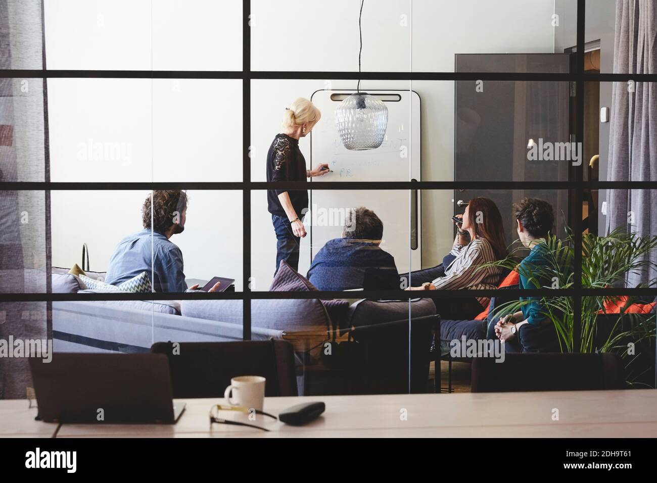 Senior femmina professionista che dà presentazione ai colleghi visti dal vetro in sala riunioni presso l'ufficio creativo Foto Stock