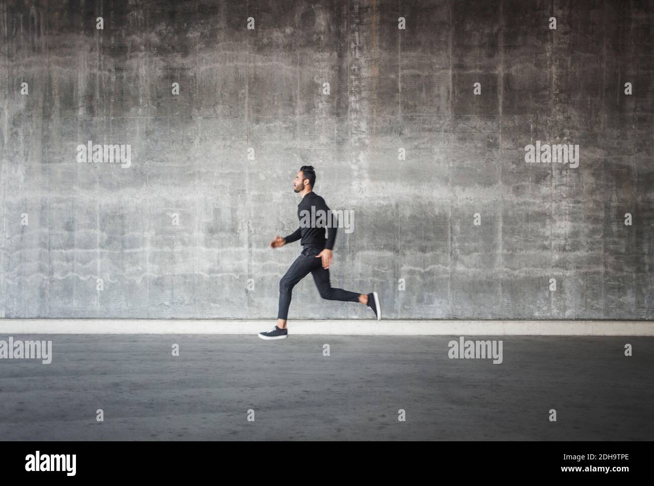 Vista laterale di un giovane uomo che corre da una parete grigia Foto Stock