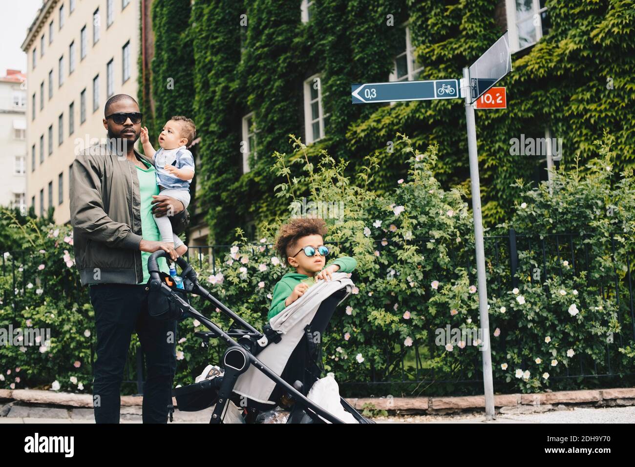 Padre che cammina con i bambini mentre spinge il passeggino del bambino contro le piante in città Foto Stock