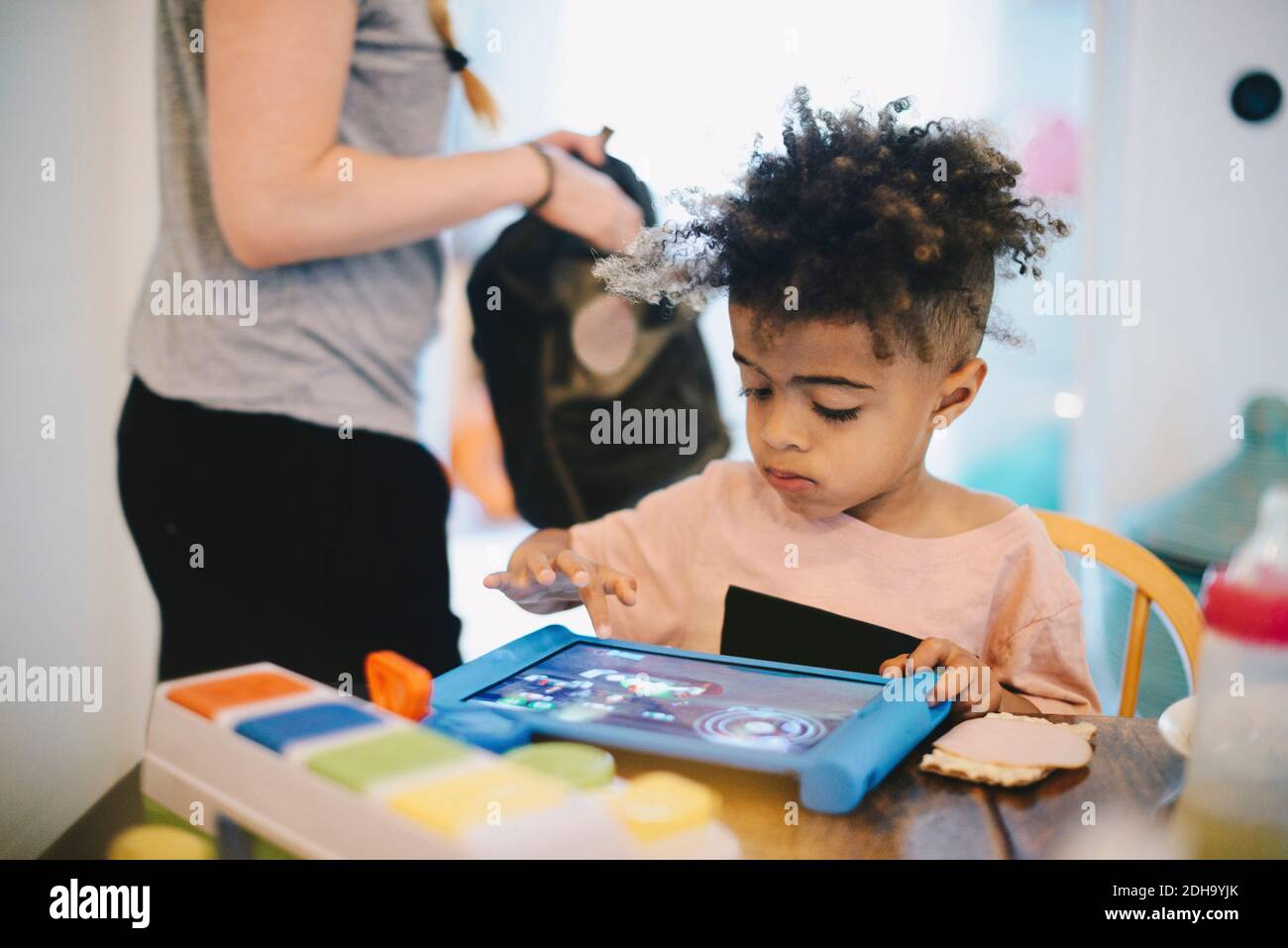 Ragazzo che usa un tablet digitale al tavolo mentre la madre si trova in piedi sfondo Foto Stock