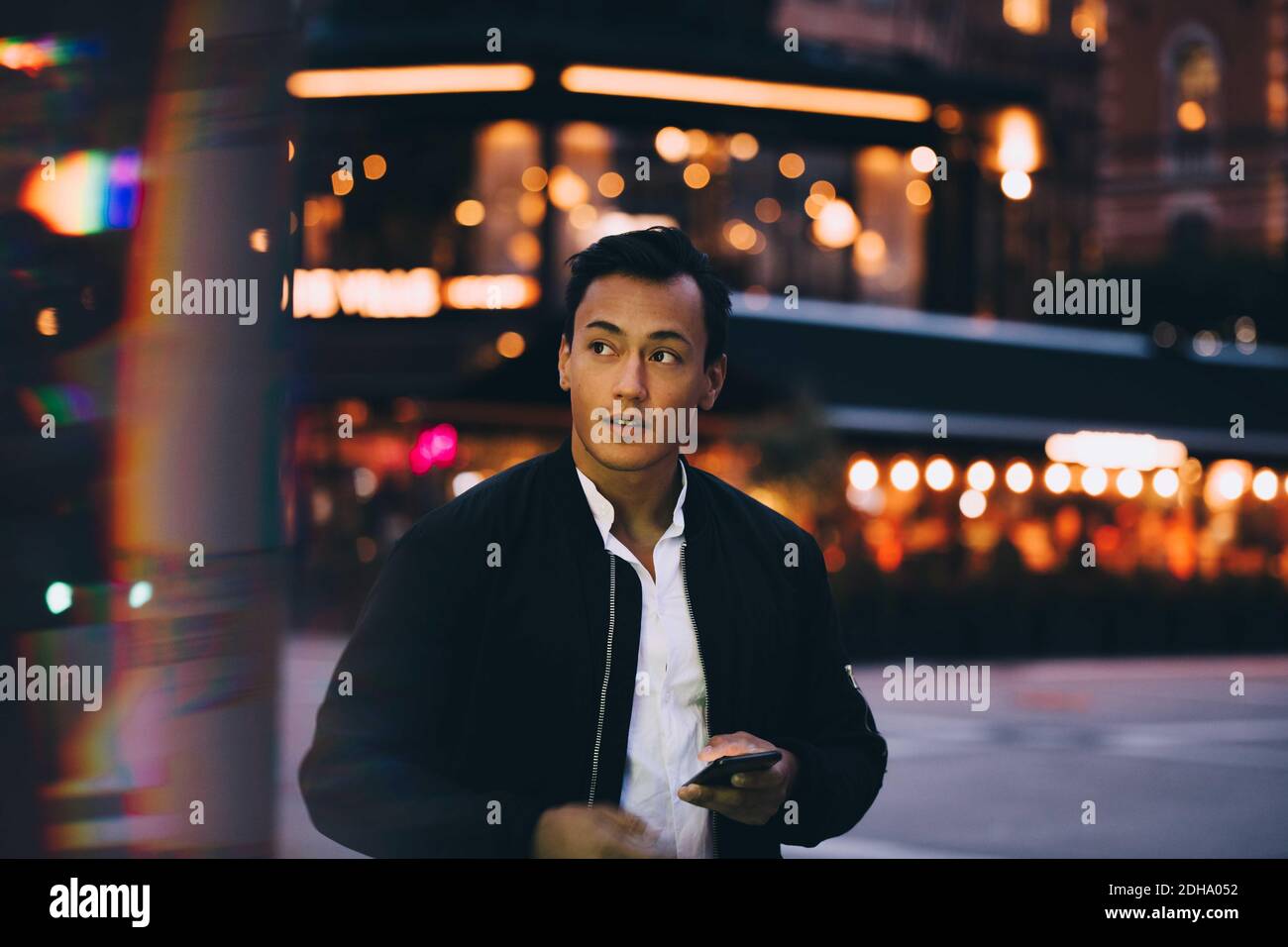 Giovane uomo con telefono che guarda lontano mentre si è in piedi illuminato città di notte Foto Stock