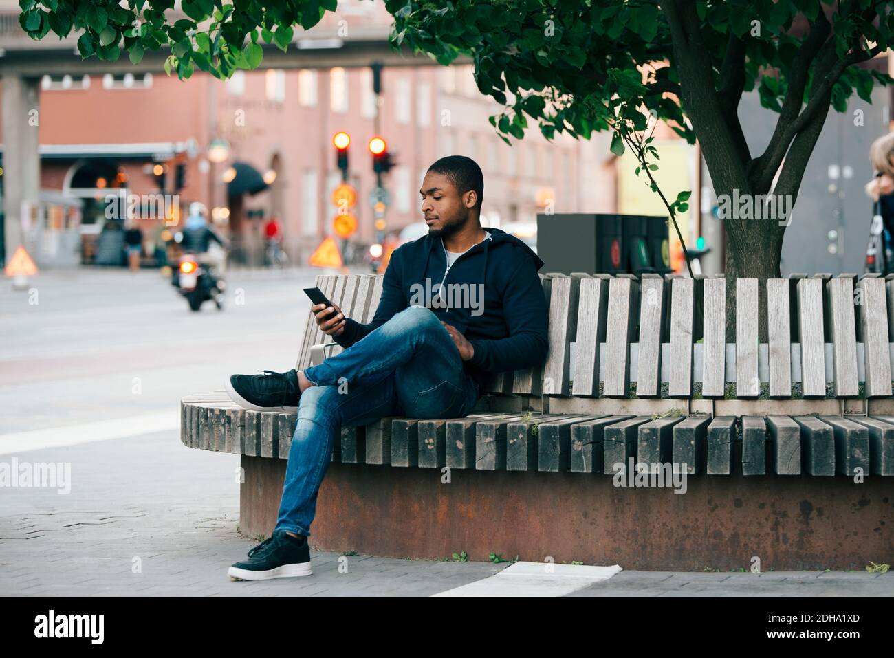 Giovane uomo che usa lo smartphone mentre si è seduti con le gambe incrociate in panchina in città Foto Stock