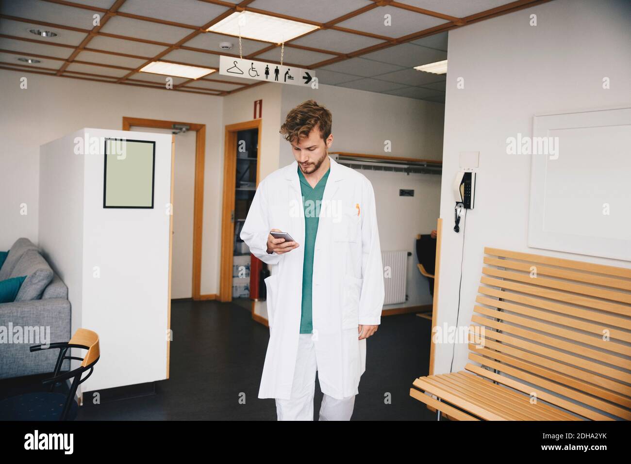 Giovane medico che usa lo smartphone mentre cammina nel corridoio a. ospedale Foto Stock