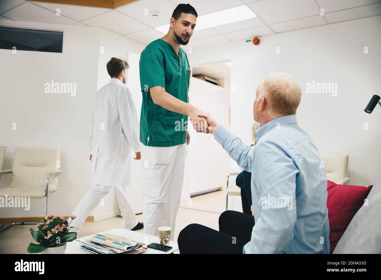 Giovane infermiere maschio che scuote le mani con paziente maturo seduto hall dell'ospedale Foto Stock