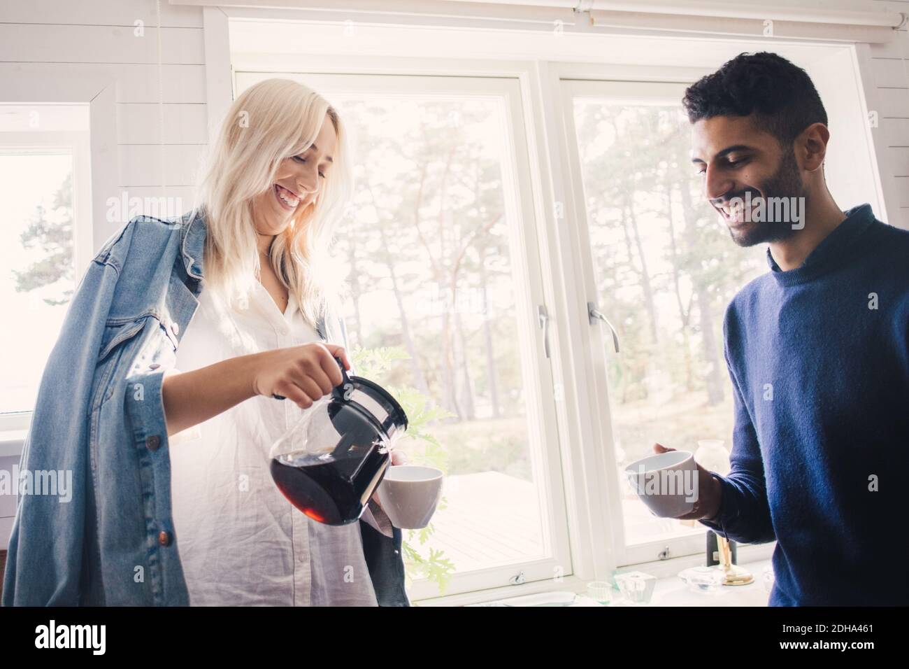 Sorridente giovane donna che versa il caffè in tazza mentre si sta in piedi amico maschile a casa Foto Stock