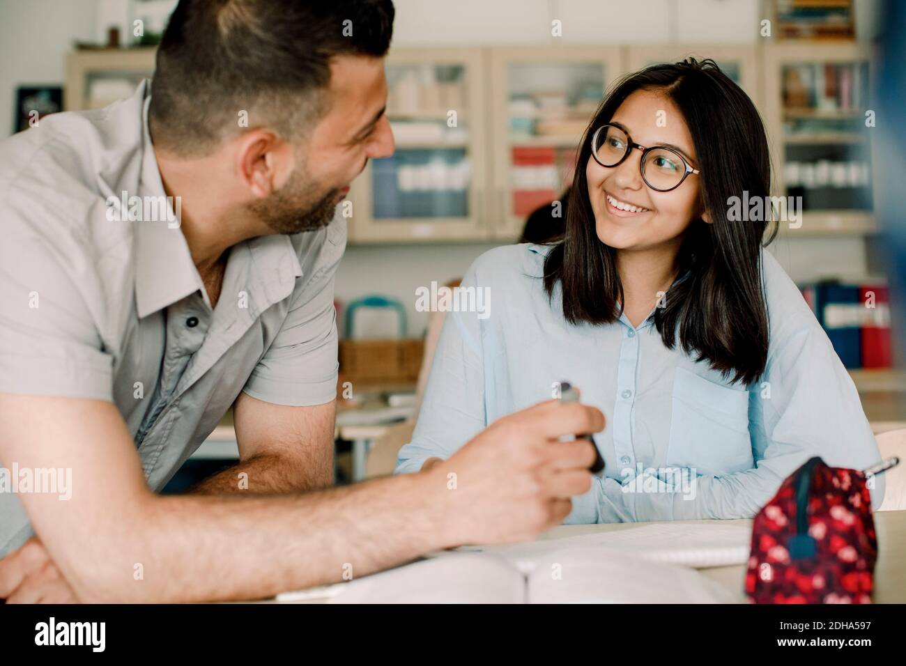 Sorridente insegnante di sesso maschile che spiega lo studente mentre si appoggia al tavolo in aula Foto Stock