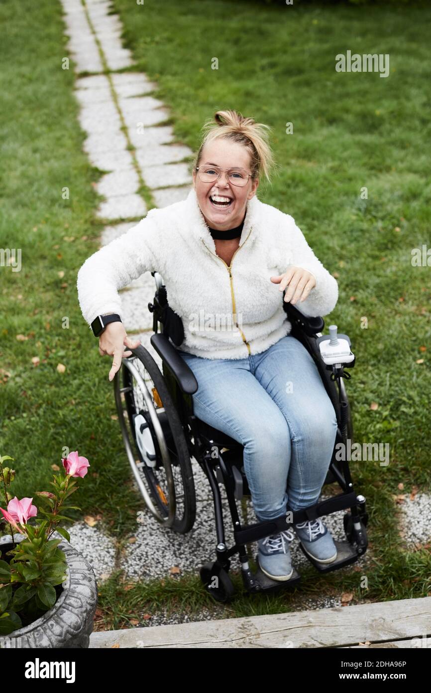 Ritratto a lunghezza intera di una donna sorridente e disabile seduta su sedia a rotelle in cortile Foto Stock