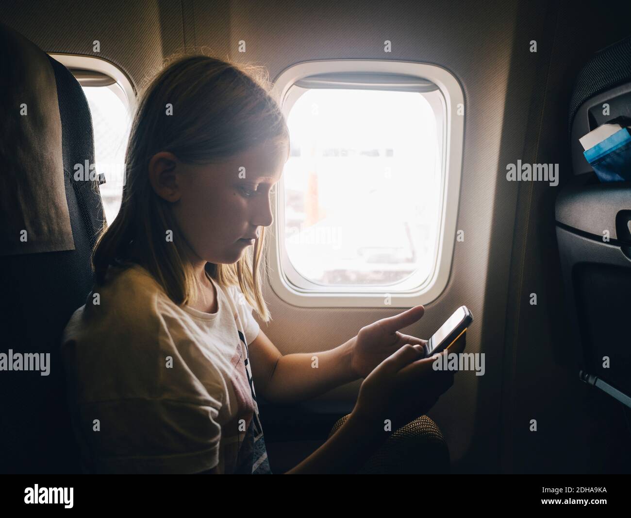 Ragazza caucasica che usa il telefono cellulare mentre si siede vicino alla finestra dell'aereo Foto Stock