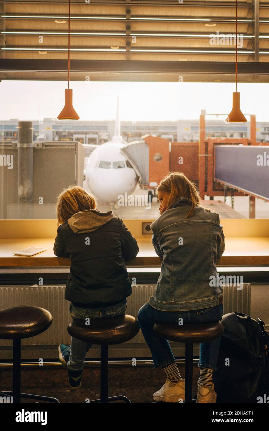 Vista posteriore di fratello e sorella seduti contro il finestrino a. aeroporto Foto Stock