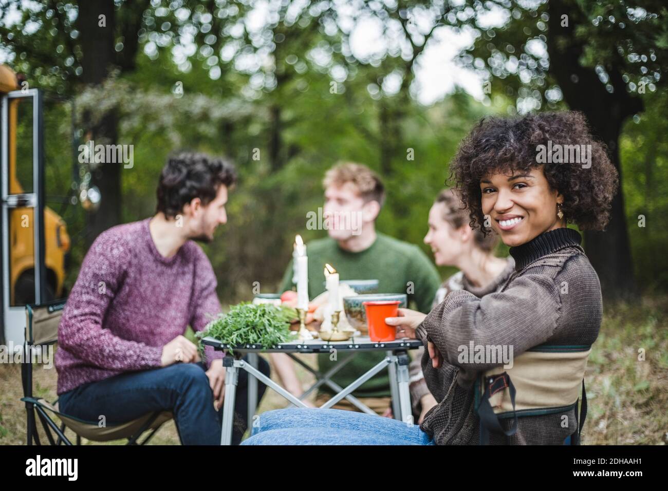 Ritratto di donna afro sorridente seduta con gli amici al tavolo durante il campeggio in foresta Foto Stock