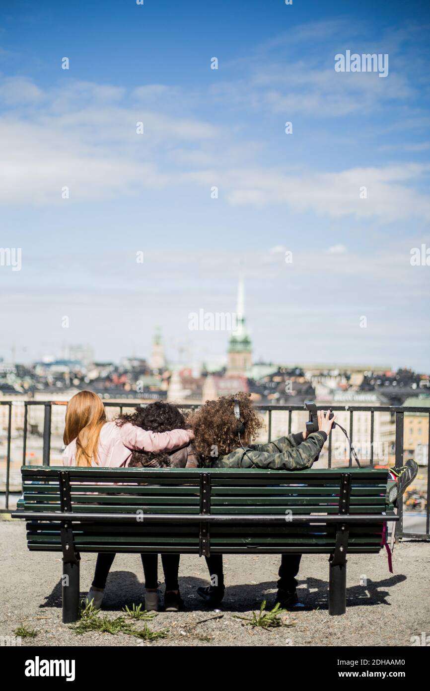 Ragazzo adolescente che prende selfie con le amiche mentre si siede sopra panca contro il cielo Foto Stock