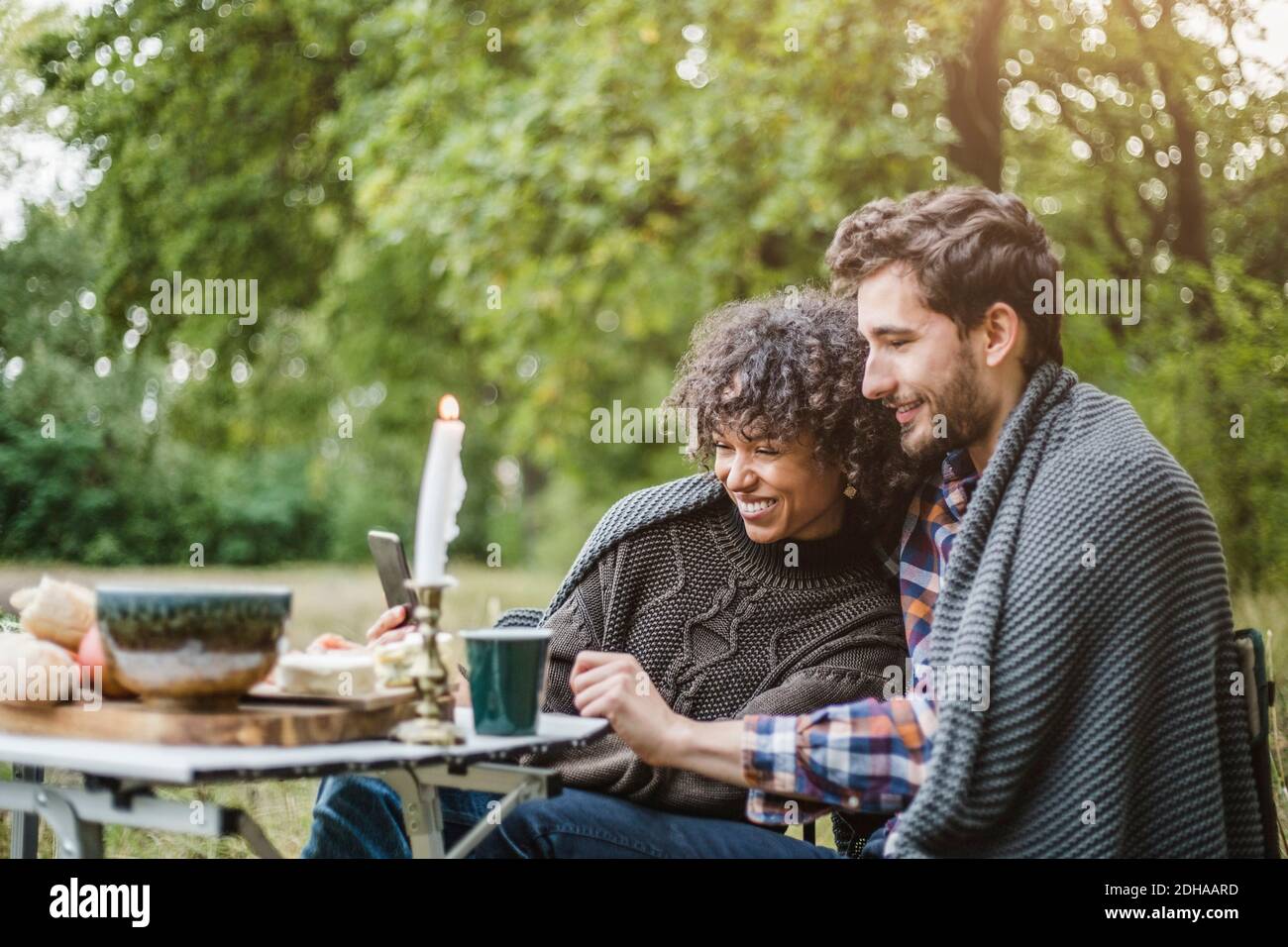 Giovane coppia allegra che condivide il telefono cellulare mentre avvolto in coperta insieme durante il campeggio in foresta Foto Stock